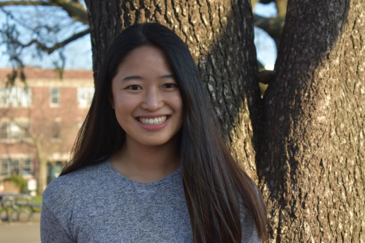 Lucy Chai, an EECS graduate student, poses in front of a campus oak. She is wearing a grey top and smiling confidently at the camera.