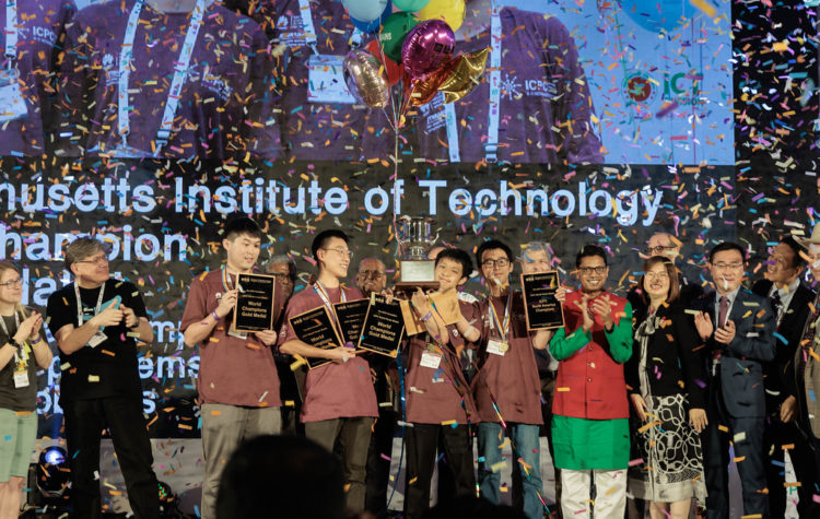 In a hail of confetti, the members of the MIT programming team stand on a large stage clutching trophies and plaques. Behind them, a huge screen displays the MIT team name.