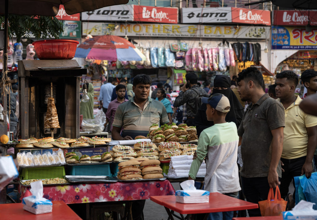 A colorful market in Dhaka features a stacked array of sandwiches and a spinning cone of meat. In the background, vendor umbrellas and packages of clothing are visible. 