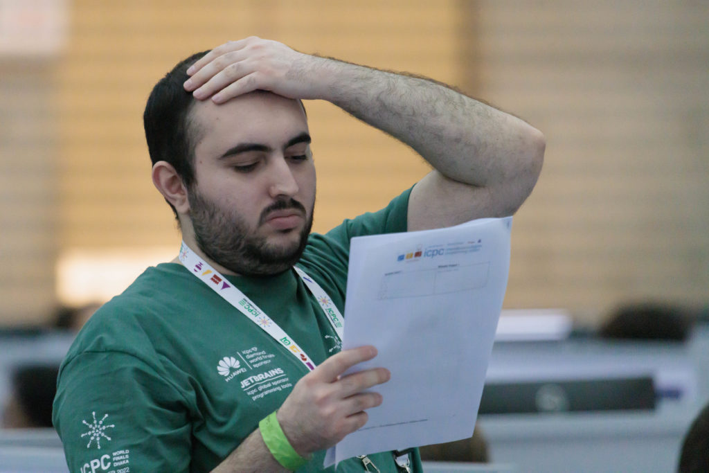 A bearded coder wearing a green t-shirt stares at a problem with a look of intense concentration. He is wiping his forehead. 