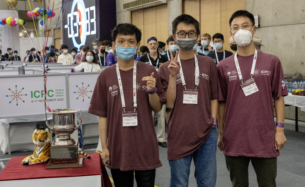 The MIT team, wearing ICPC badges, face masks, and matching burgundy t-shirts, pose next to a large trophy cup and a small plush tiger toy.