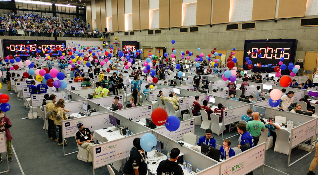 A crowded hall full of low cubicles is livened up with hundreds of colorful balloons and several huge timekeeping clocks. Inside each cubicle, a team of 3 computer programmers sit, wearing colorful t-shirts that identify their university. 