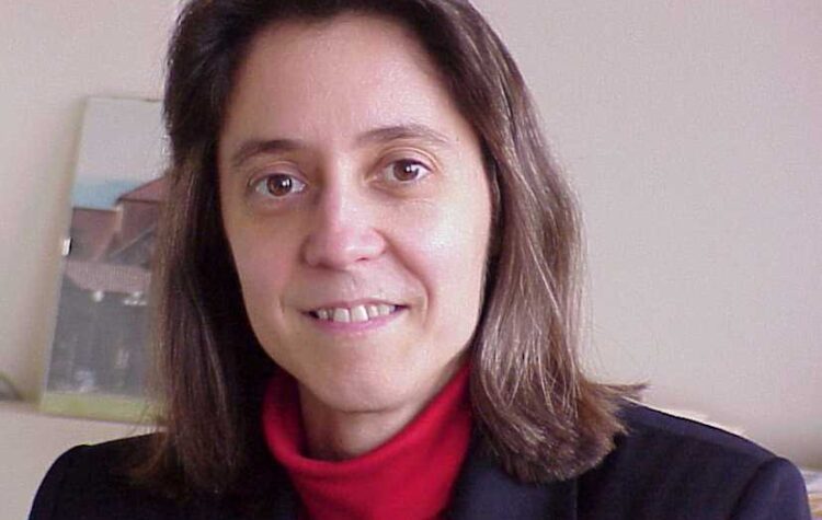 Judy Hoyt, seen here in her office in front of a photo of Stanford's campus, wears a red turtleneck and black blazer.