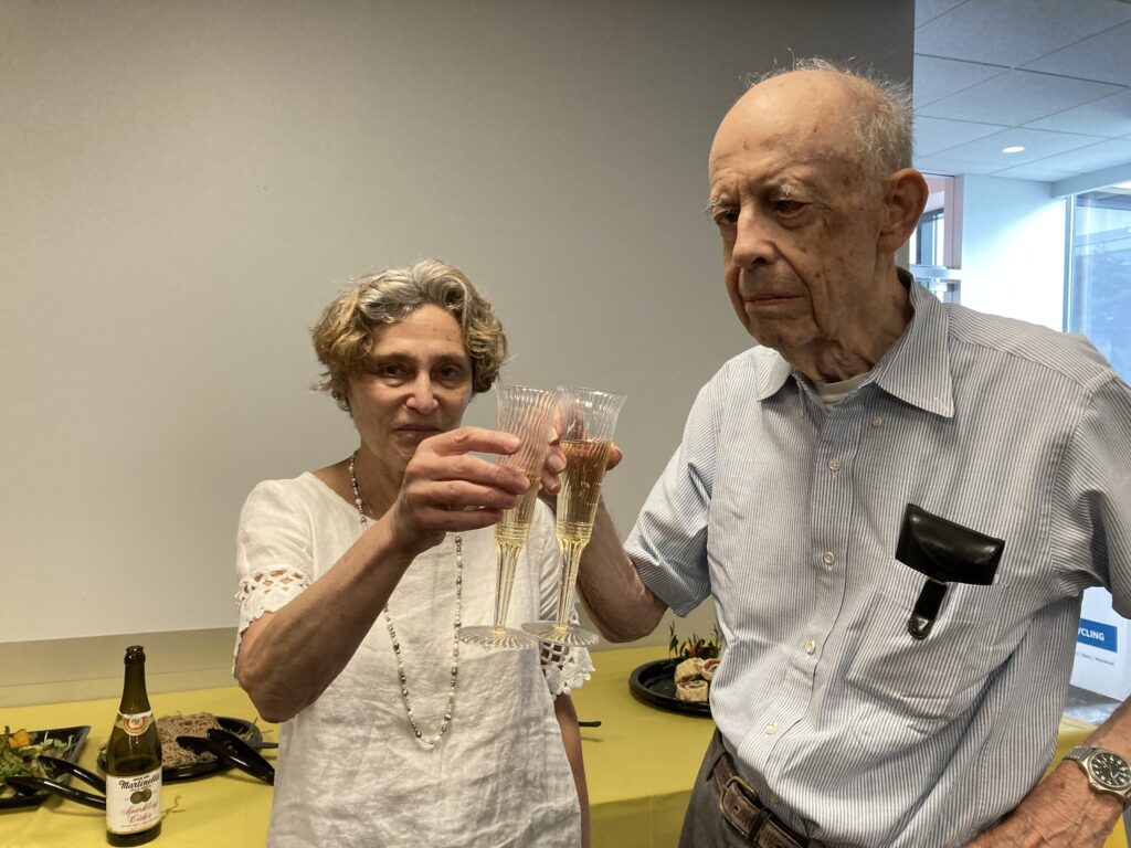 Helen Schwartz, a lady wearing a lace top and beaded necklace, toasts Fred Hennie, a man wearing a striped shirt and pocket protector. Both wear expressions of pride. 