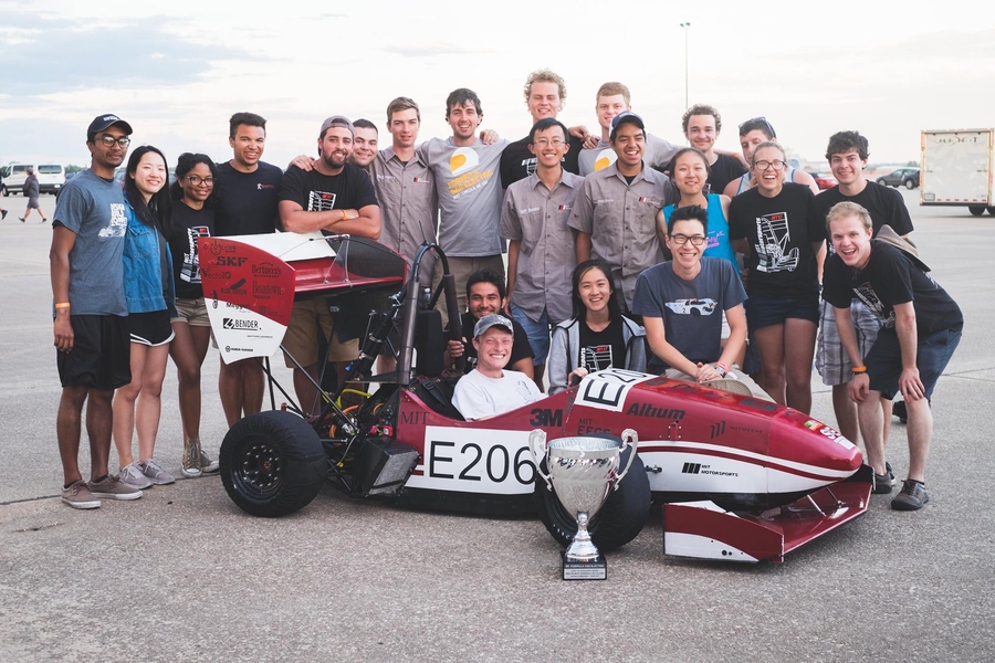 A diverse team of college students gather around a red and white racecar with a high rear spoiler. In the foreground, a silver cup sits on the asphalt.