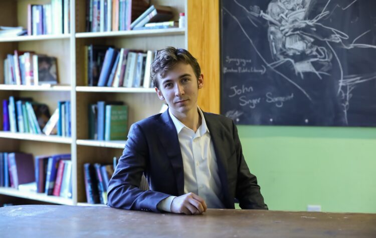 Carson, a young man with light brown hair and a serious expression, sits wearing a suit jacket and white dress shirt. He is in a lounge containing bookshelves and a messily erased blackboard.
