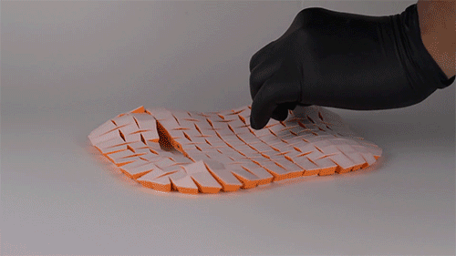 As two hands pull a string, a connected grid of small, square plastic tiles is reshaped into a softly curving structure, much like a pillow.